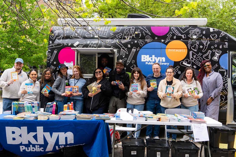 A group of individuals holding books in front of a mobile library with a vibrant design showcasing the 'Bklyn Public Library' logo.