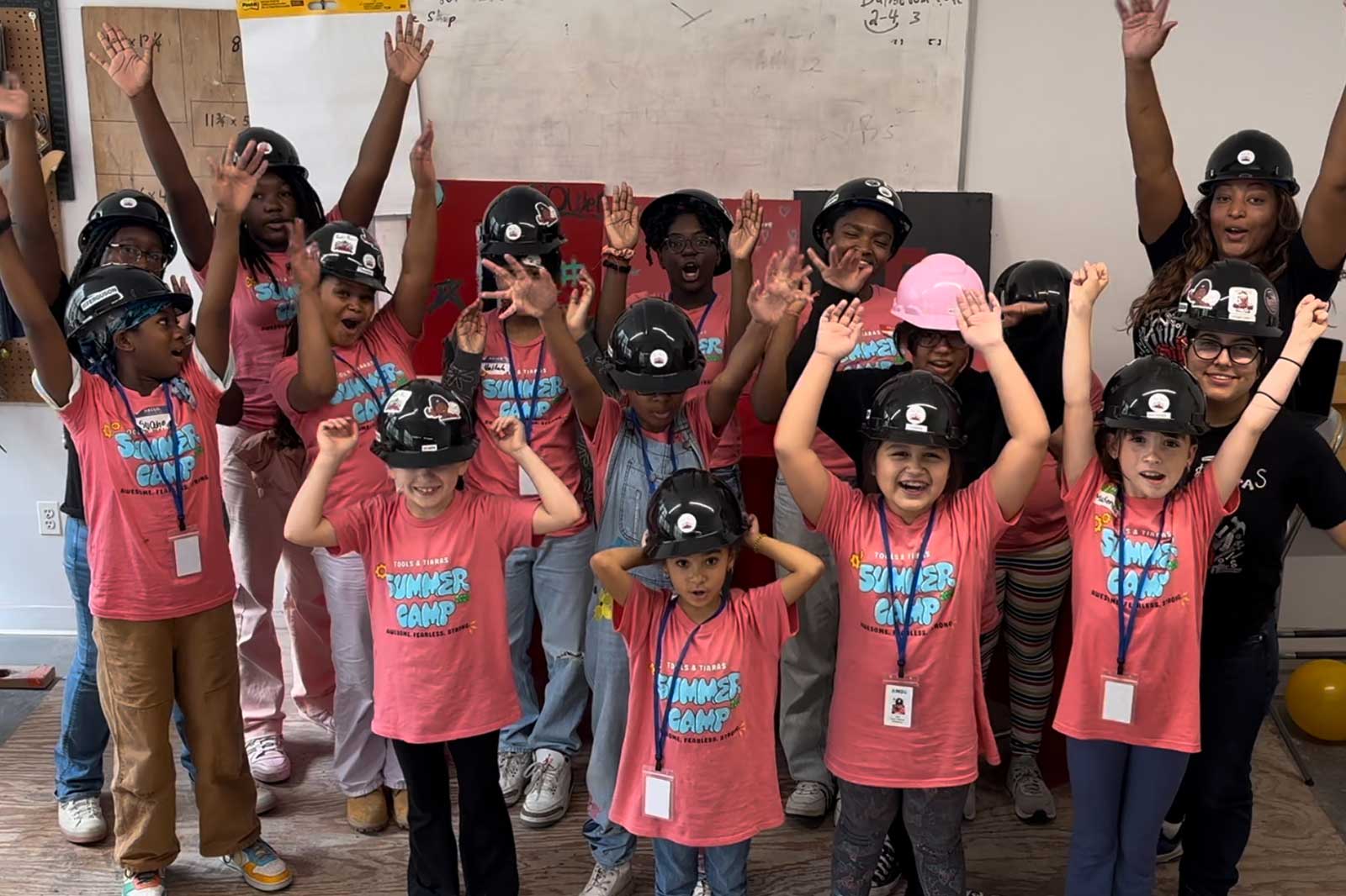 A group of children in pink t-shirts with 'SUMMER CAMP' raising their hands. They wear helmets and name tags.