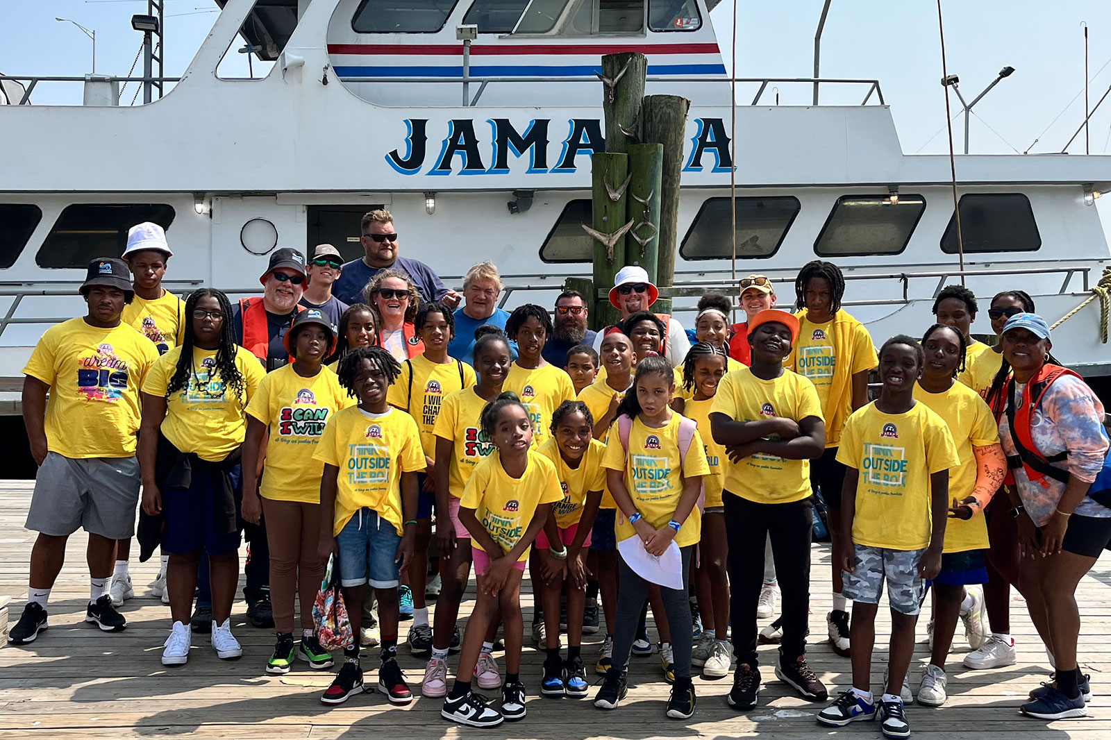 A group of children in yellow T-shirts poses on a pier with a boat named 'JAMAICA' in the background.