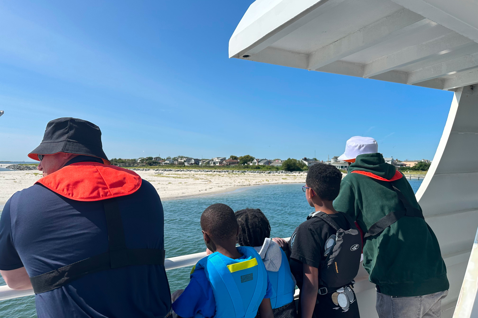 A group of children and adults watch the shoreline from a boat under a clear blue sky.