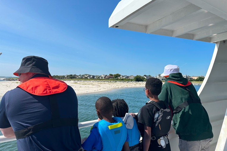 A group of children and adults watch the shoreline from a boat under a clear blue sky.