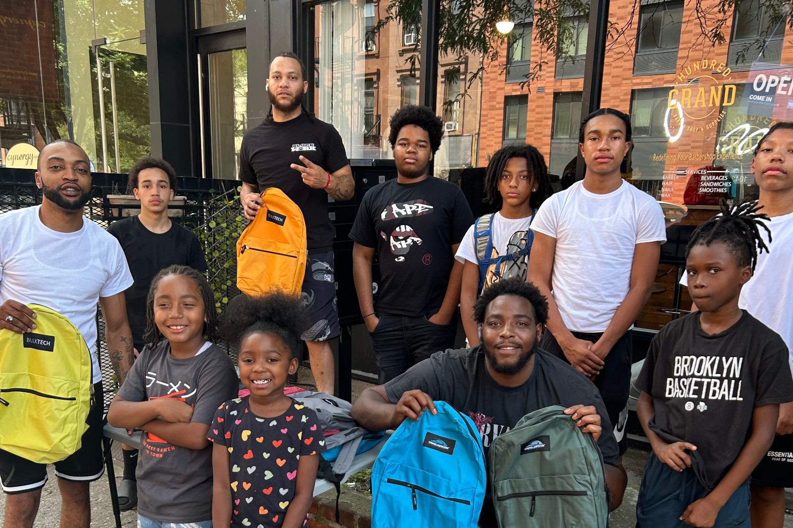 A group of individuals with backpacks in varied colours, posing outside a café named 'Hundred Grand'.