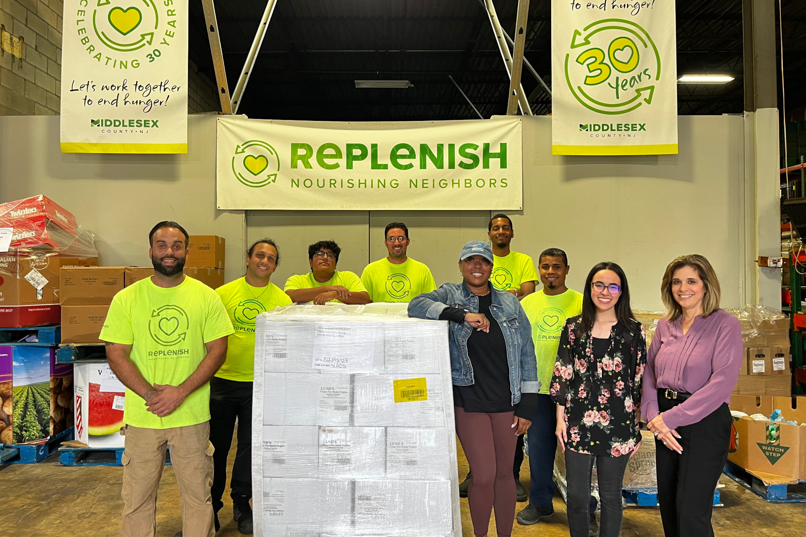 A group of volunteers in bright yellow shirts stands in front of a food donation warehouse, promoting the Replenish initiative.