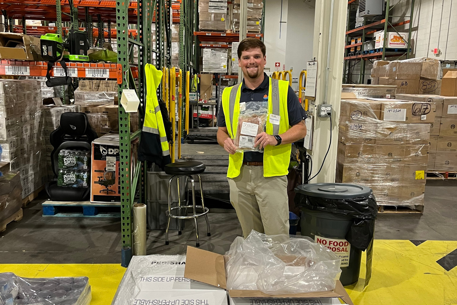 A man in a safety vest holds a package in a warehouse filled with stacked boxes and pallets.