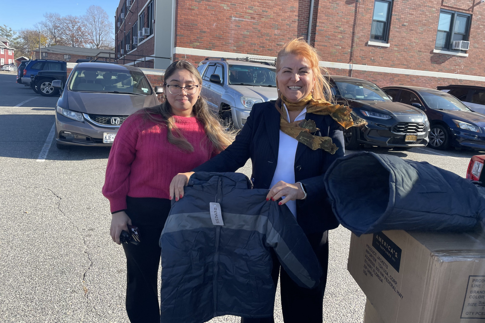Two women hold a grey jacket in a parking lot, surrounded by parked cars and a brown brick building.