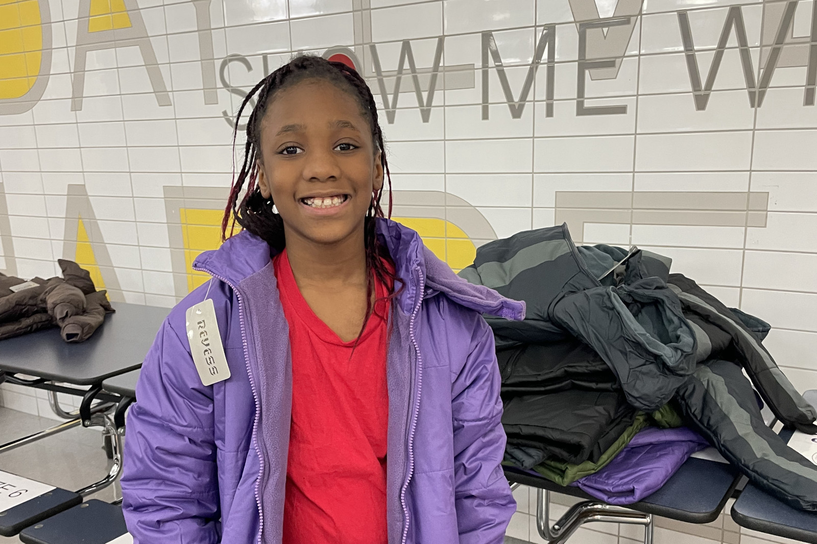 A child in a purple jacket and red shirt stands by a pile of jackets in a tiled room with the phrase 'SHOW ME WHO' on the wall.