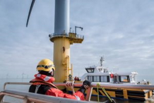 A crew member wearing a helmet and life jacket prepares to board a service boat beside a wind turbine in a calm sea.