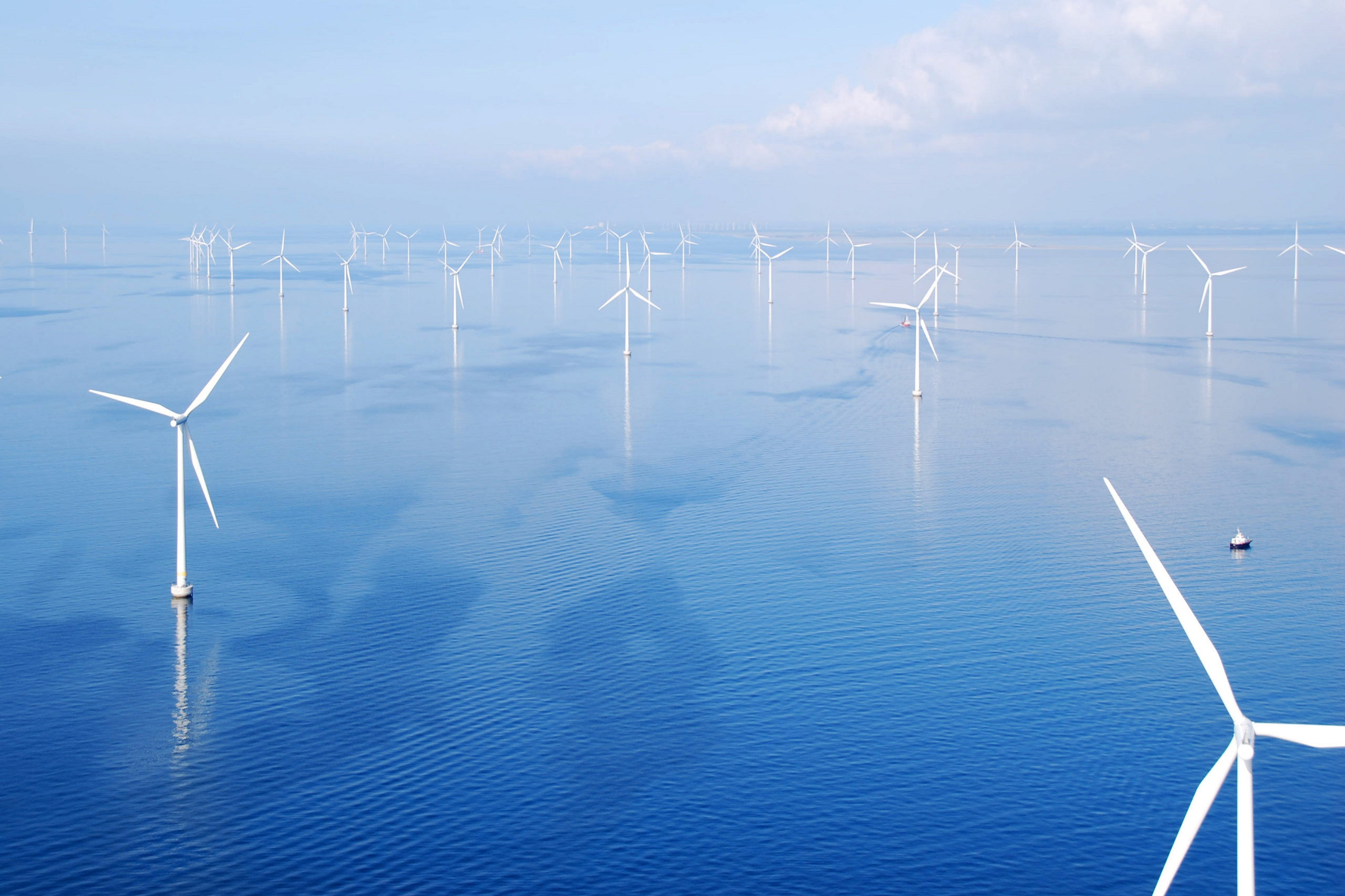 A panoramic view of numerous white wind turbines in a blue ocean under a clear sky, with a small boat in the distance.