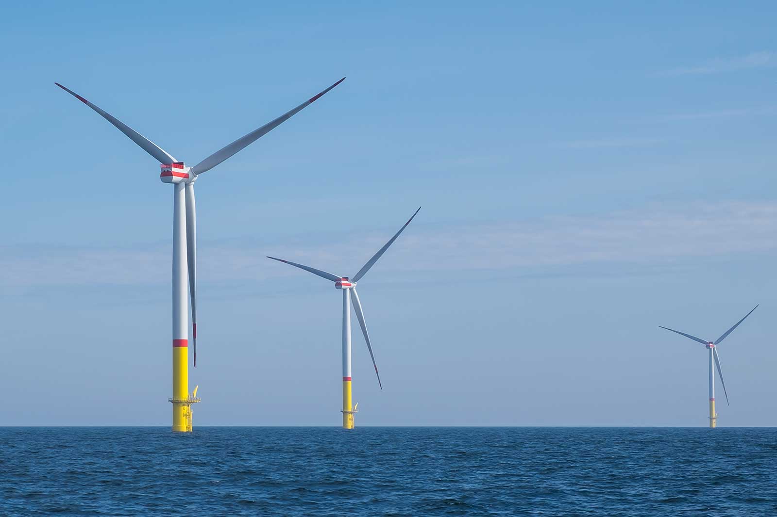 Three wind turbines stand in calm waters under a clear blue sky, with colourful bases and blades spinning gracefully.